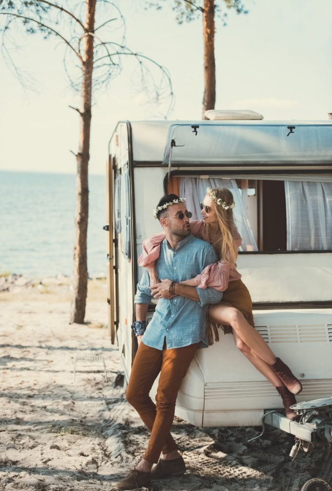 hippie couple in wreaths and sunglasses embracing and sitting on trailer